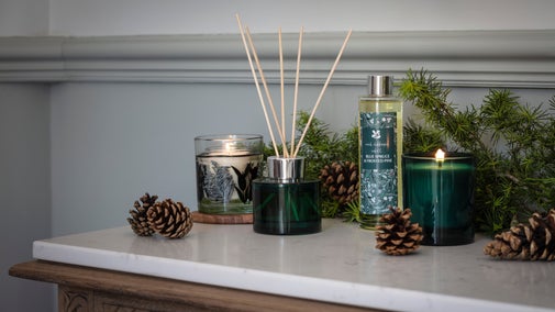 Autumn home fragrance collection with candles, reed diffuser, pine cones and evergreen foliage on a marble-topped table.
